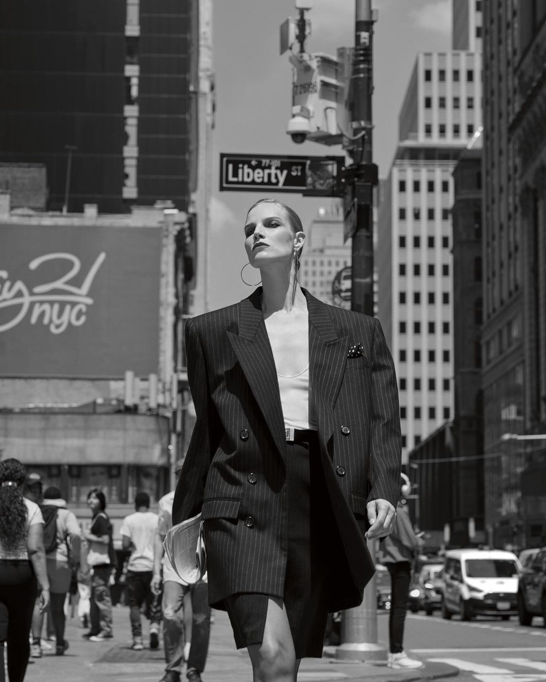 Fashionable adult woman in an oversized pinstripe blazer and skirt walks on a city street with pedestrians and cars.