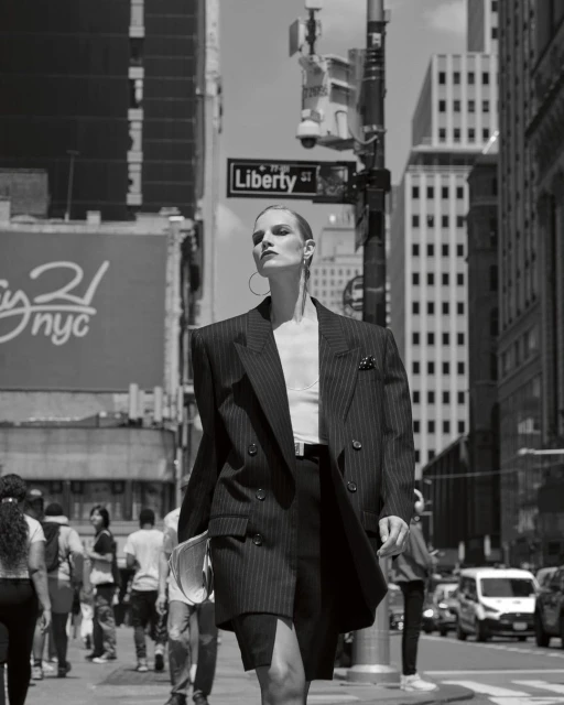 Fashionable adult woman in an oversized pinstripe blazer and skirt walks on a city street with pedestrians and cars.