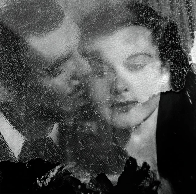 Monochrome image of a man and woman's faces behind a cracked glass surface, creating a distorted, artistic effect.