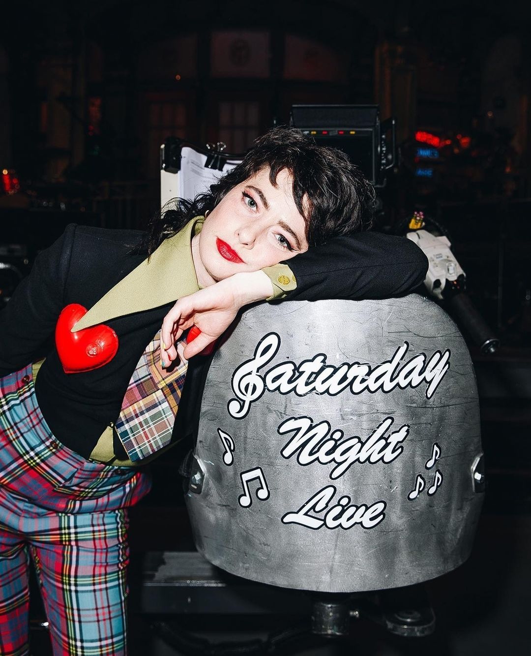 Adult female rests head on Saturday Night Live prop; wearing red heart brooch, plaid pants and tie; medium, eye-level shot.