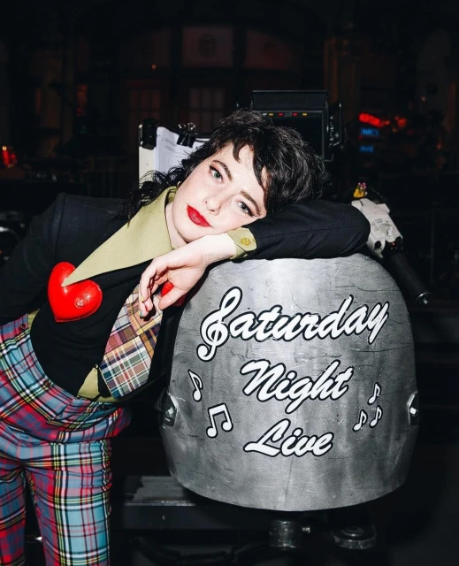 Adult female rests head on Saturday Night Live prop; wearing red heart brooch, plaid pants and tie; medium, eye-level shot.
