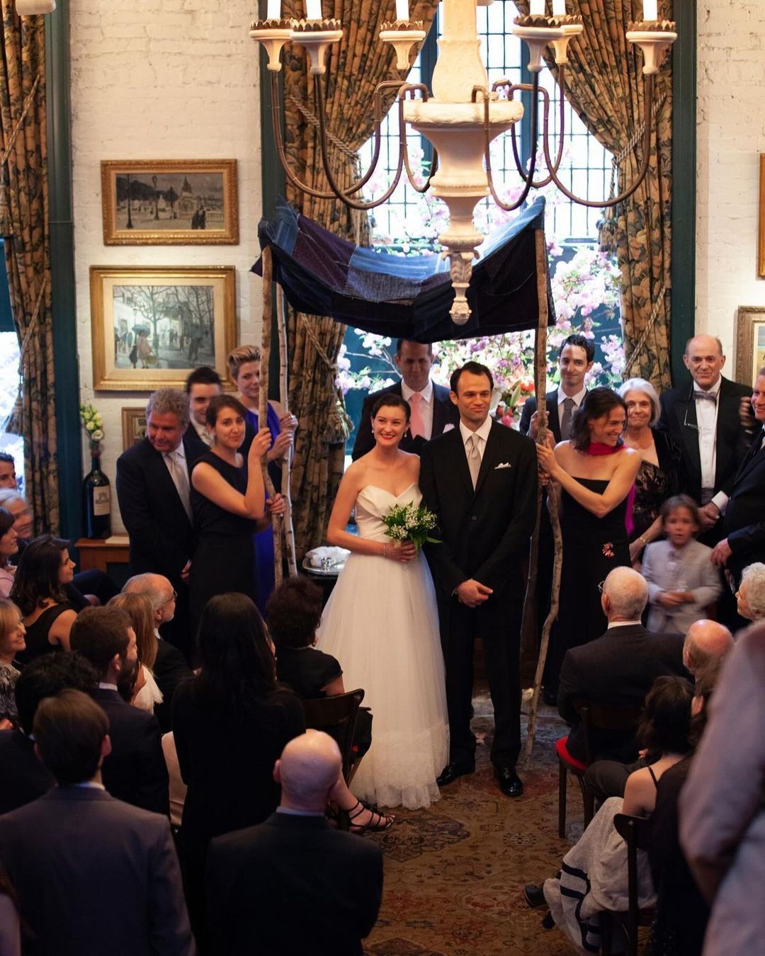 Indoor wedding ceremony with bride and groom under a chuppah, surrounded by guests in formal attire.