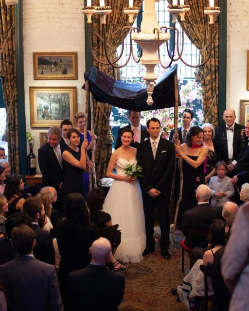 Indoor wedding ceremony with bride and groom under a chuppah, surrounded by guests in formal attire.