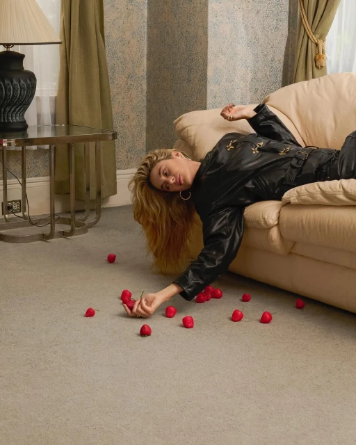 A blonde woman in a black leather jumpsuit reclines on a beige couch, surrounded by red berries scattered on a grey carpet indoors.