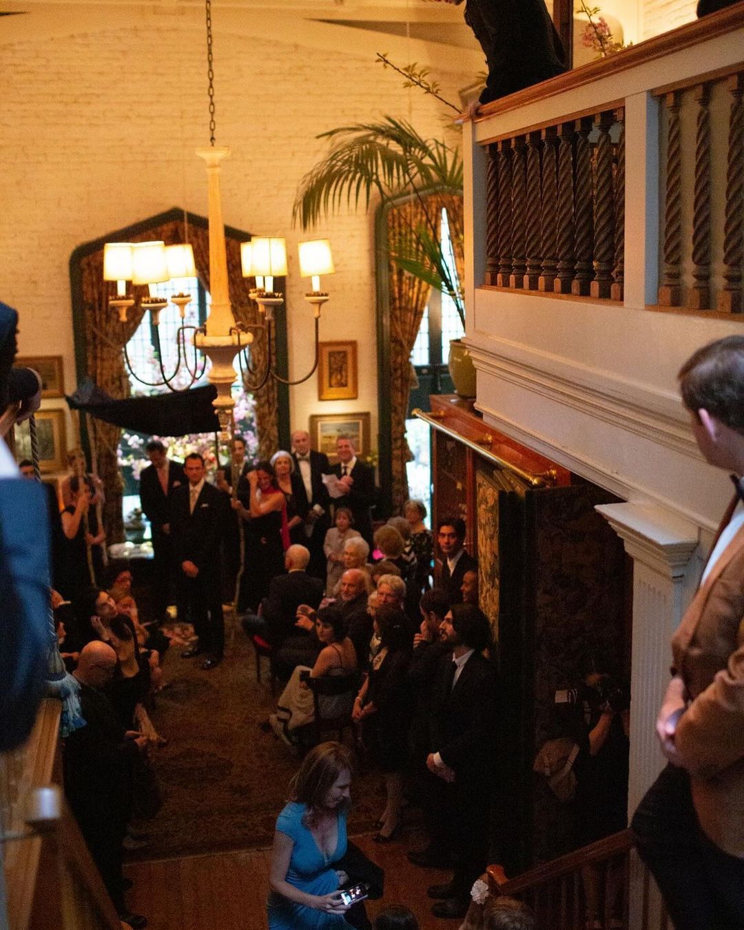Large group of people, some in suits and dresses, gather in an indoor space with a winding staircase and high ceilings for an event.