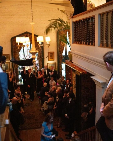 Large group of people, some in suits and dresses, gather in an indoor space with a winding staircase and high ceilings for an event.