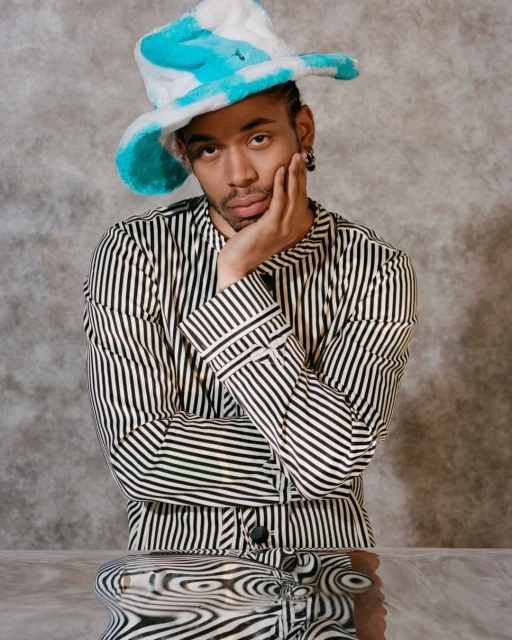 Stylish person poses with a striped top and a white and blue hat in an indoor studio setting, complemented by a reflective table.