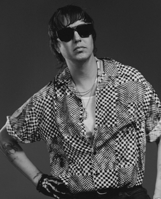 Monochrome portrait of an adult male wearing sunglasses and a checkered shirt, posing indoors against a dark grey background.