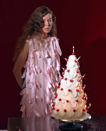 Woman in pale pink dress adorned with ribbons standing beside a tall multi-tiered frosted cake with lit candles and cherries.