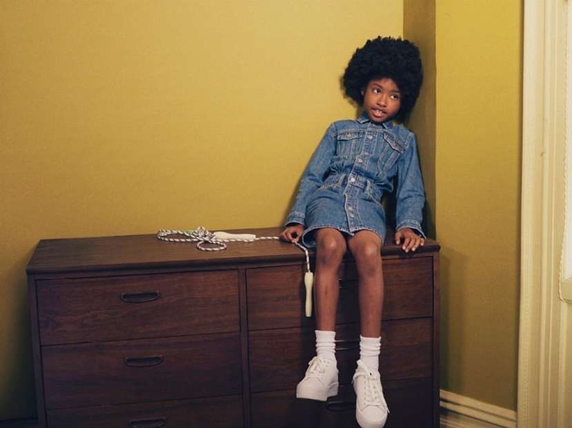 Young girl with an afro in a denim dress sits on a wooden dresser with a jump rope in an indoor room with a yellow wall.