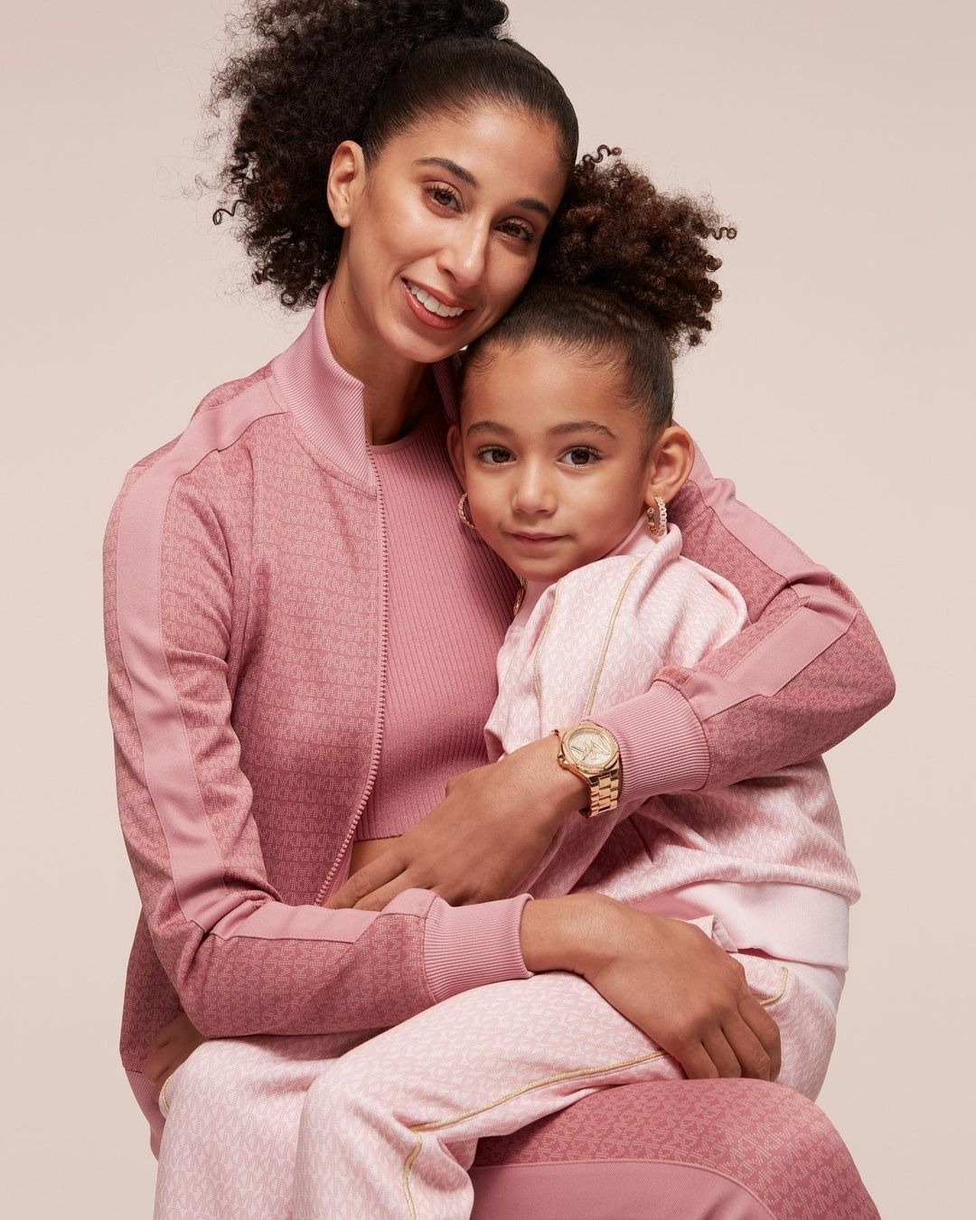 Mother and daughter in matching pink tracksuits pose indoors against a neutral background. Close-up studio shot with soft lighting.