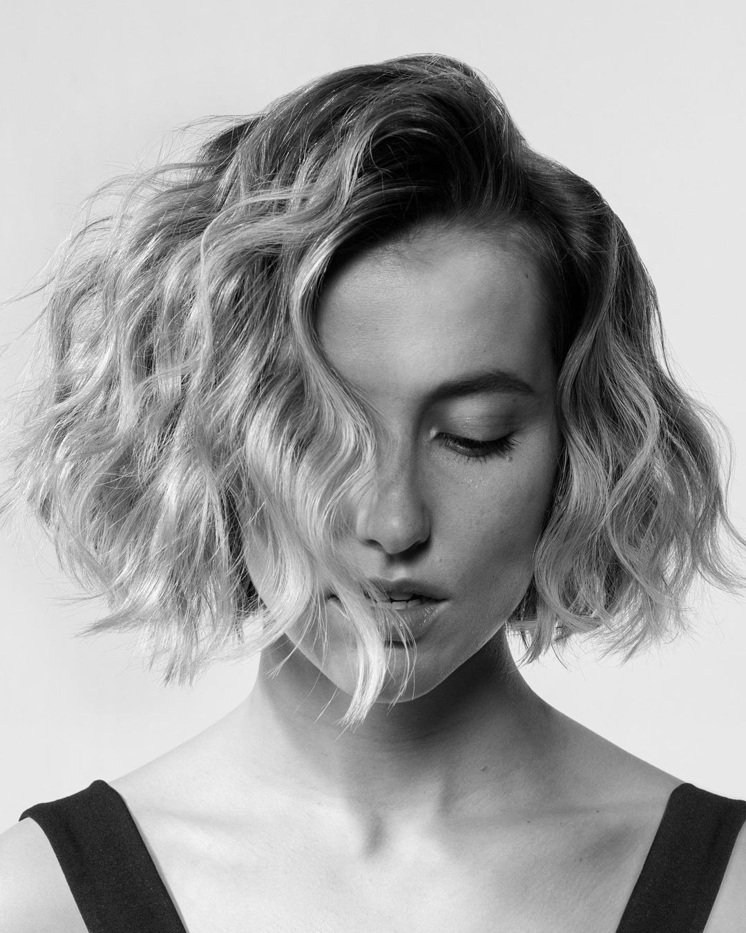 Monochrome portrait of a young adult woman with wavy blonde bob, closed eyes, and black top, against a neutral background.