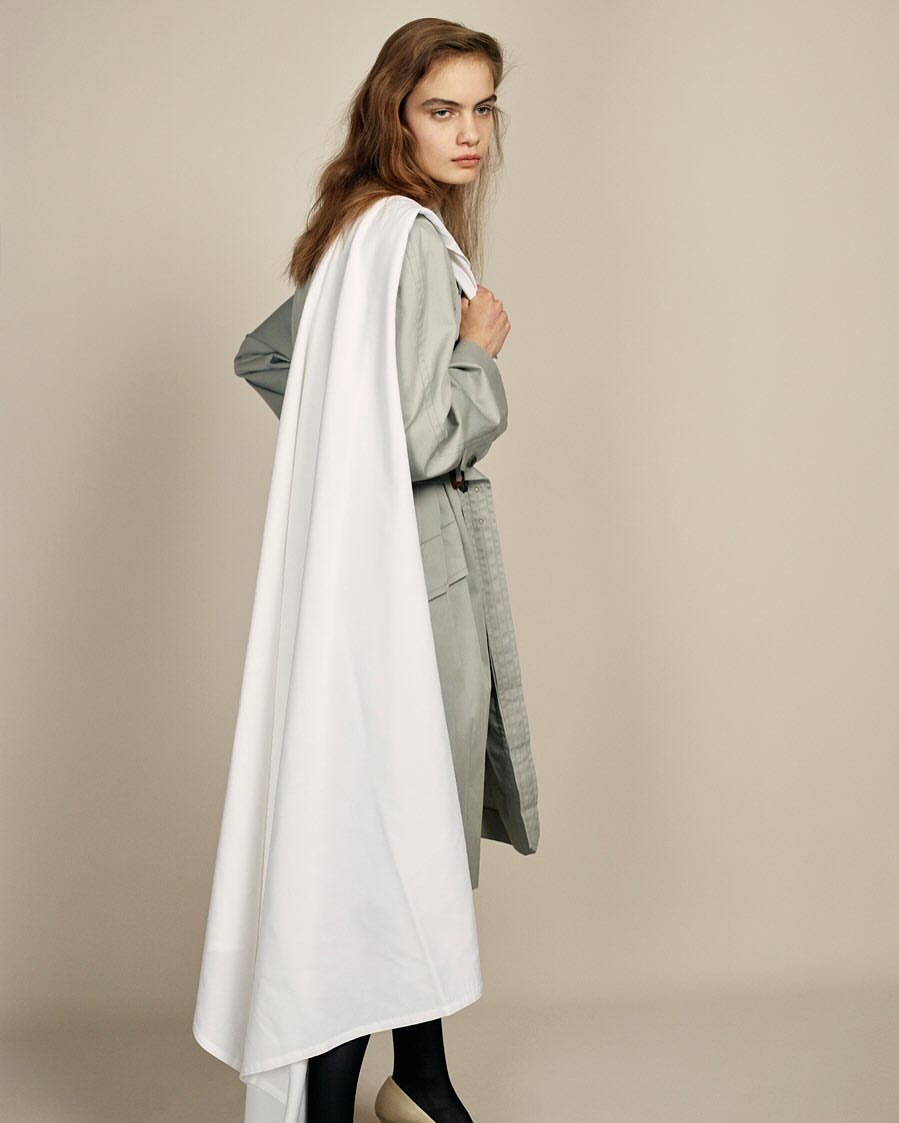 Elegant young woman in a trendy belted trench coat and black stockings stands gracefully in a fashion studio setting, looking stylish.