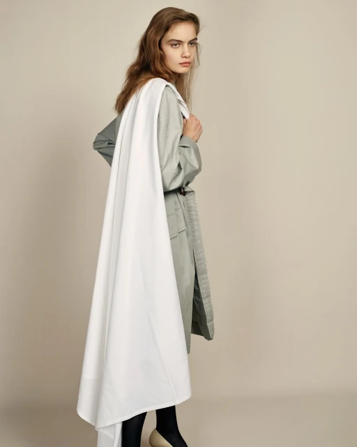 Elegant young woman in a trendy belted trench coat and black stockings stands gracefully in a fashion studio setting, looking stylish.