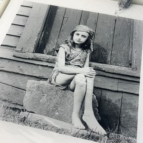 Monochrome portrait of a barefoot child in costume with headscarf sitting on a stone step in front of a weathered wooden building.