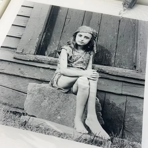 Monochrome portrait of a barefoot child in costume with headscarf sitting on a stone step in front of a weathered wooden building.