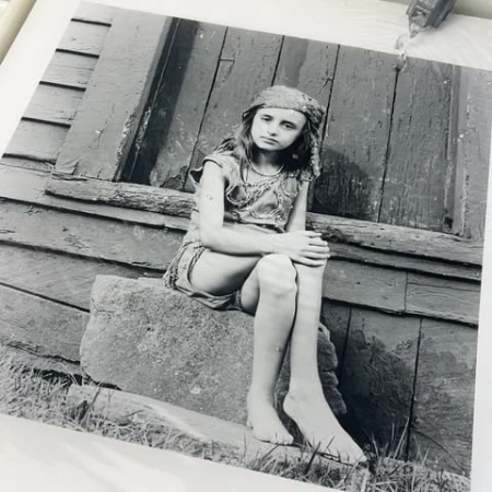 Monochrome portrait of a barefoot child in costume with headscarf sitting on a stone step in front of a weathered wooden building.