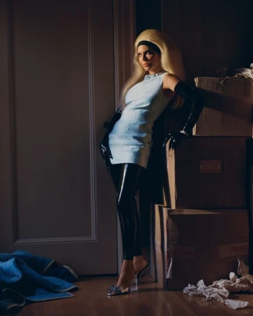 Model with voluminous blonde hair wearing a light blue minidress and black leggings, posing beside stacked cardboard boxes in a room.