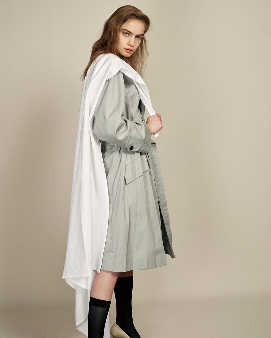Young woman poses in a light grey trench coat and white cape, paired with black socks and tan heels, against a neutral backdrop.