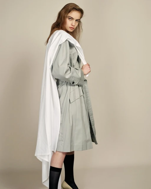 Young woman poses in a light grey trench coat and white cape, paired with black socks and tan heels, against a neutral backdrop.