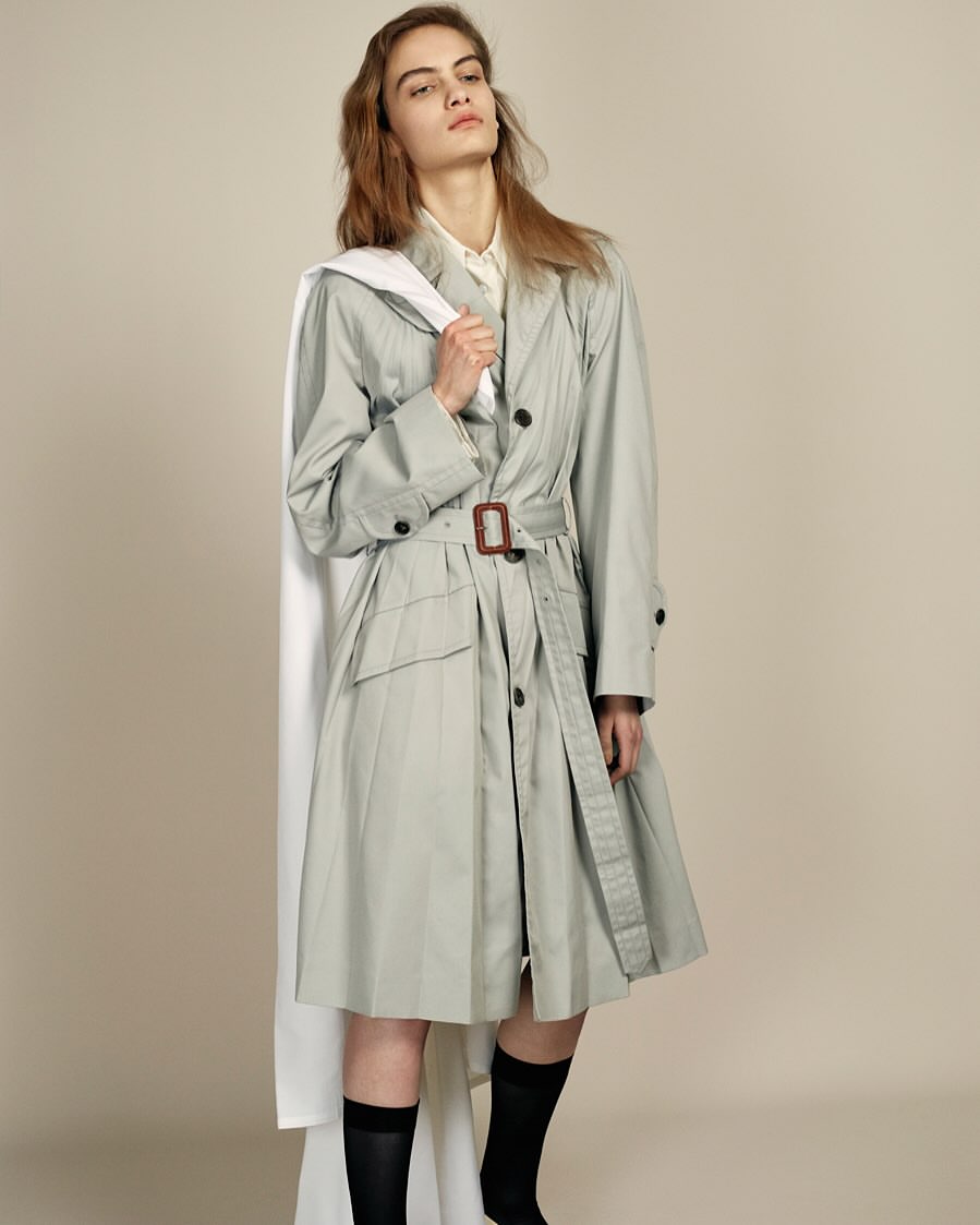 Fashionable adult female poses in a belted trench coat with black socks, holding a white garment bag in a light-filled studio.