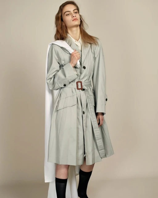 Fashionable adult female poses in a belted trench coat with black socks, holding a white garment bag in a light-filled studio.