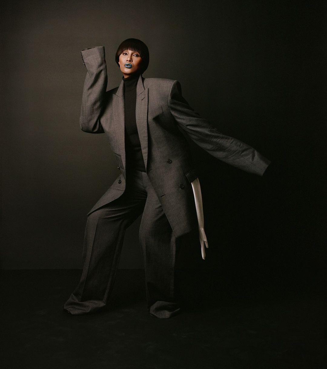 Adult female poses in oversized grey suit with sculptural silhouette against a dark backdrop in a fashion-forward studio shot.