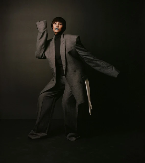 Adult female poses in oversized grey suit with sculptural silhouette against a dark backdrop in a fashion-forward studio shot.