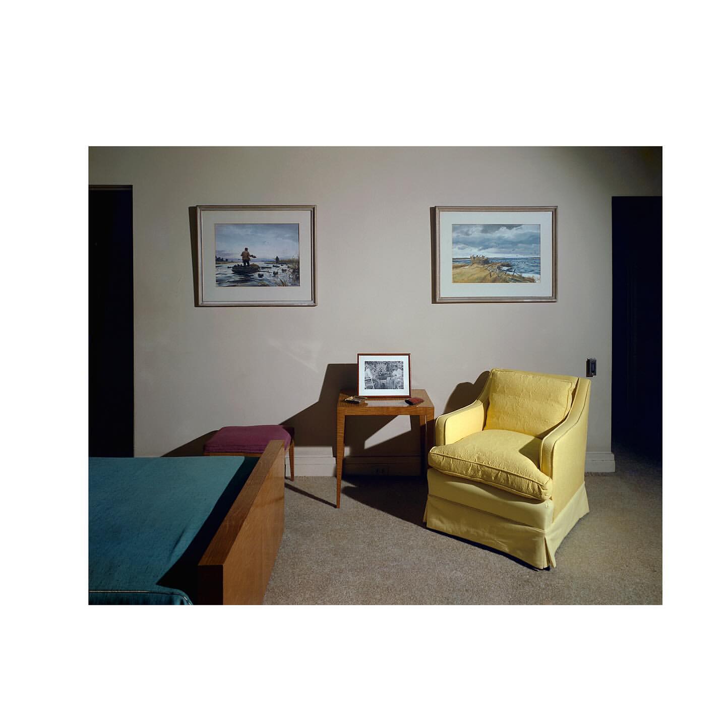 Living room interior featuring a yellow armchair, side table with framed picture, artwork, and a bed with teal bedding in a neutral setting.