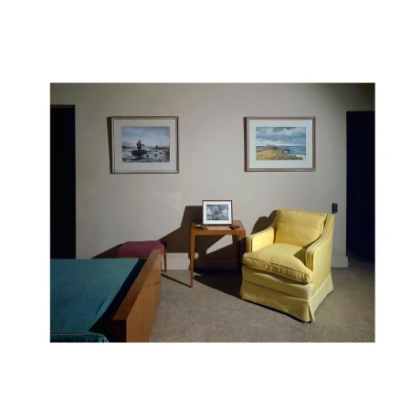 Living room interior featuring a yellow armchair, side table with framed picture, artwork, and a bed with teal bedding in a neutral setting.