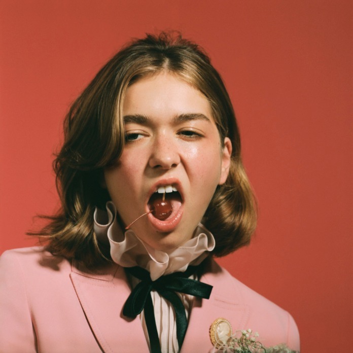 Portrait of a young adult woman with a cherry in her mouth, wearing a pink suit, against a red background in a photo studio.