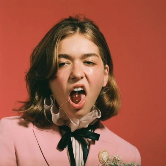 Portrait of a young adult woman with a cherry in her mouth, wearing a pink suit, against a red background in a photo studio.