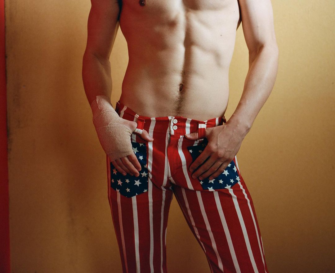 Torso of a shirtless adult male wearing red-and-white striped pants with star pockets, set against a tan and red wall.