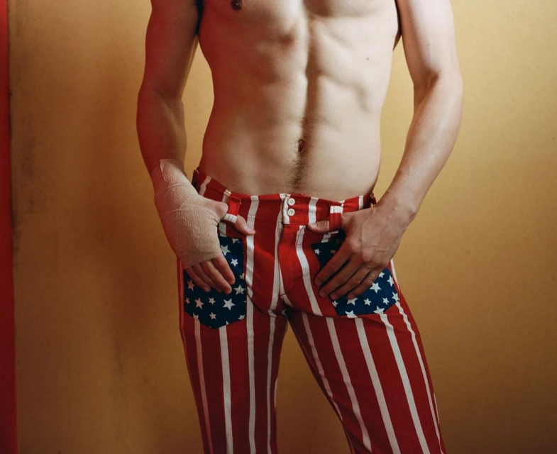 Torso of a shirtless adult male wearing red-and-white striped pants with star pockets, set against a tan and red wall.