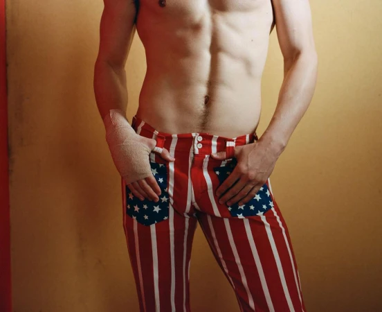 Torso of a shirtless adult male wearing red-and-white striped pants with star pockets, set against a tan and red wall.