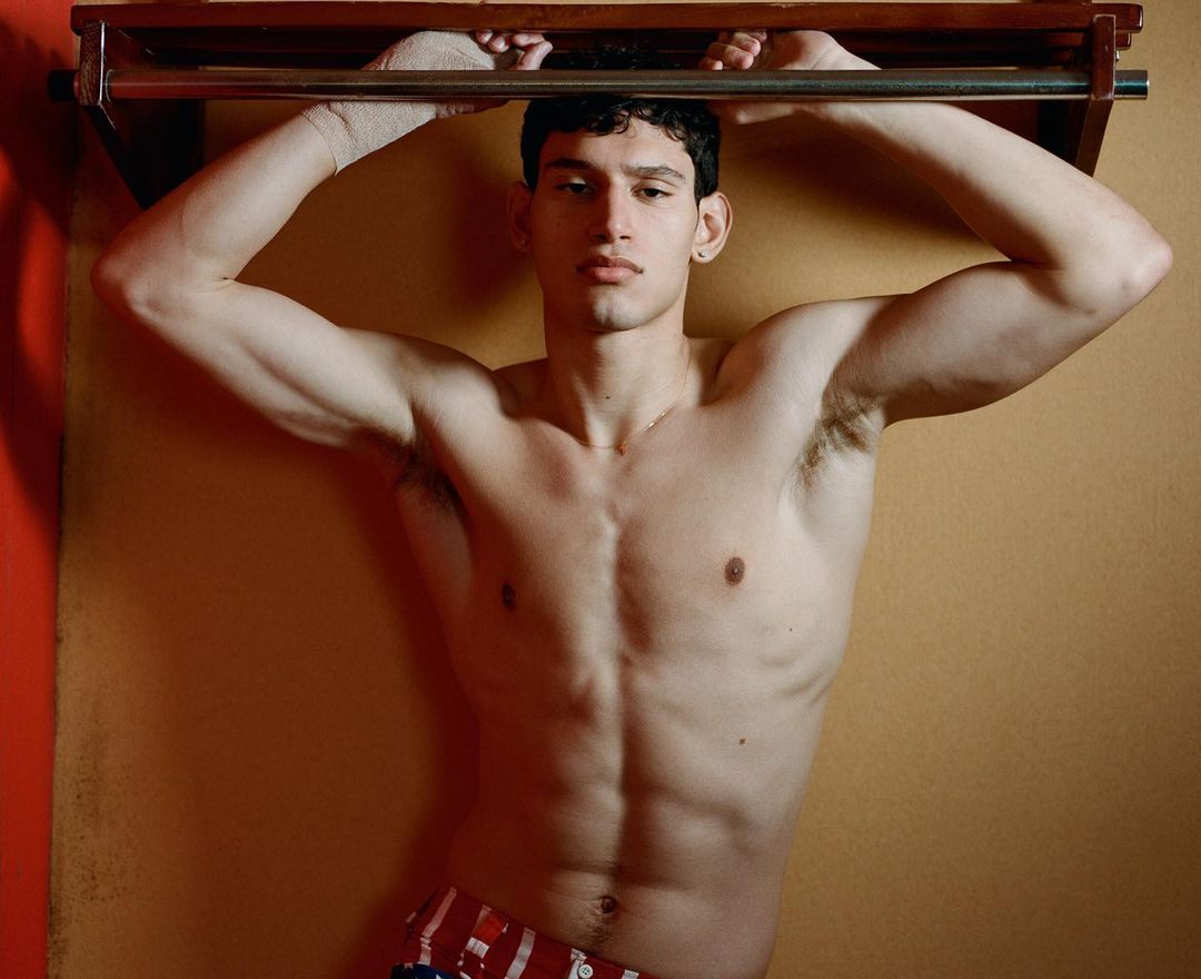 Shirtless adult male poses indoors while gripping a metal bar; he is fair-skinned with dark hair in a medium shot against a neutral wall.