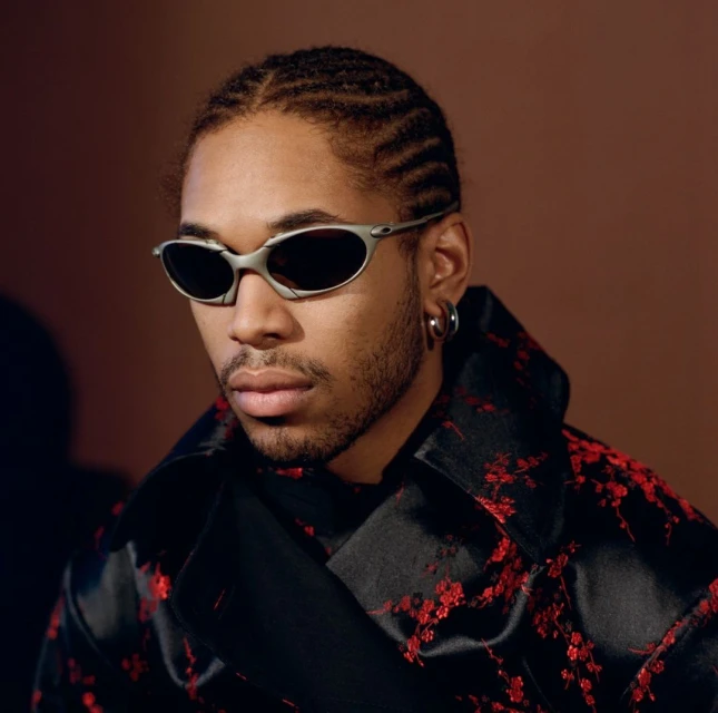 Portrait of a young adult male with cornrow braids, wearing sunglasses and a black jacket with a red floral pattern.