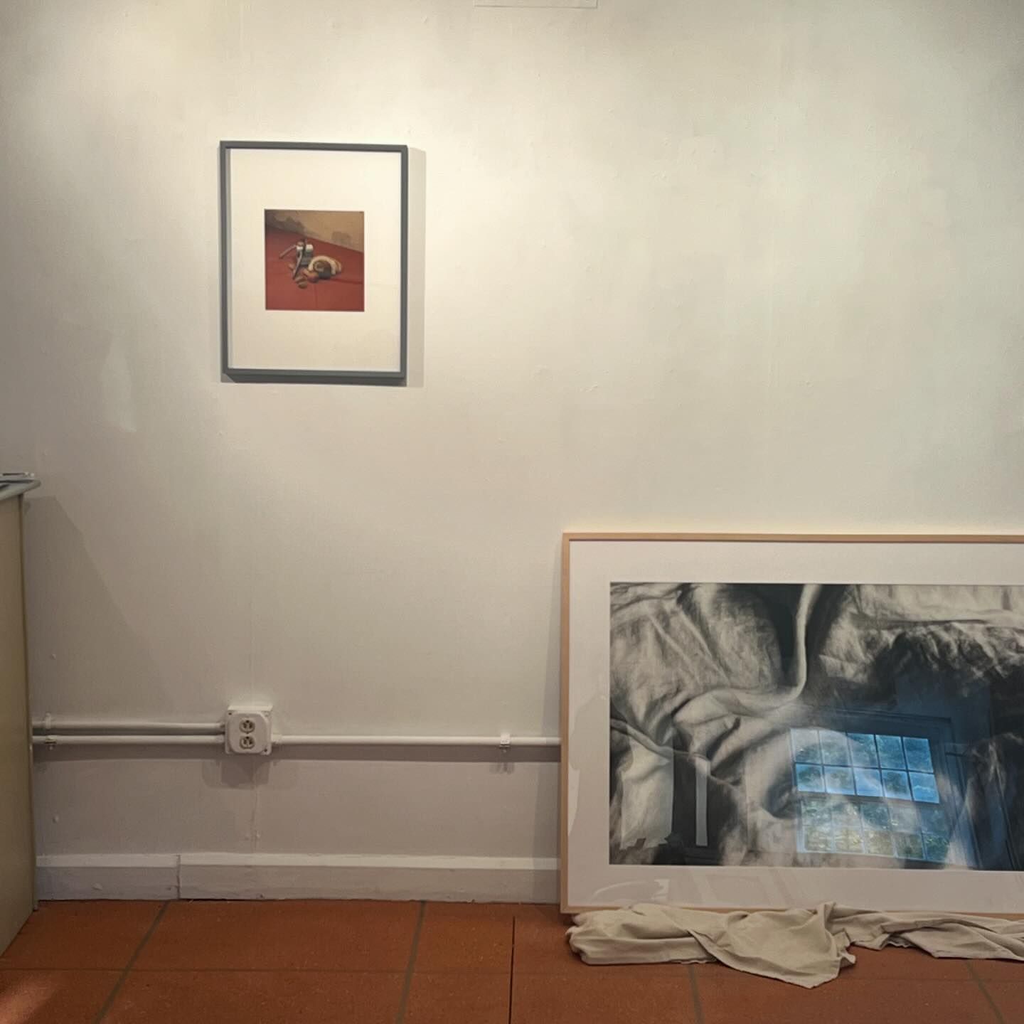 Interior view of an art gallery displaying two framed artworks on a white wall, with a tiled floor adding a sense of depth and perspective.
