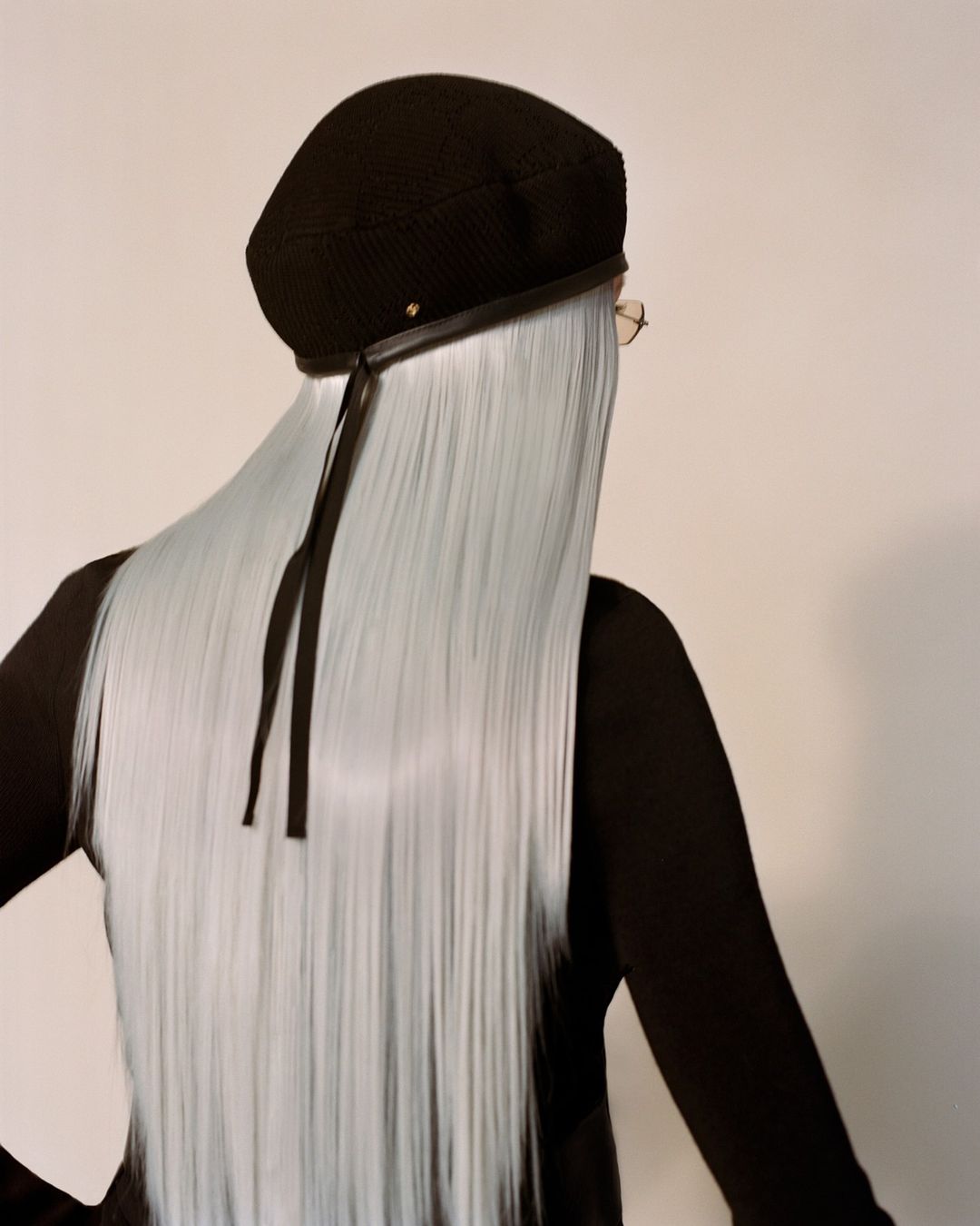 Fashionable adult female with long blue hair wears a black beret with ribbons, back view, studio setting, neutral background.