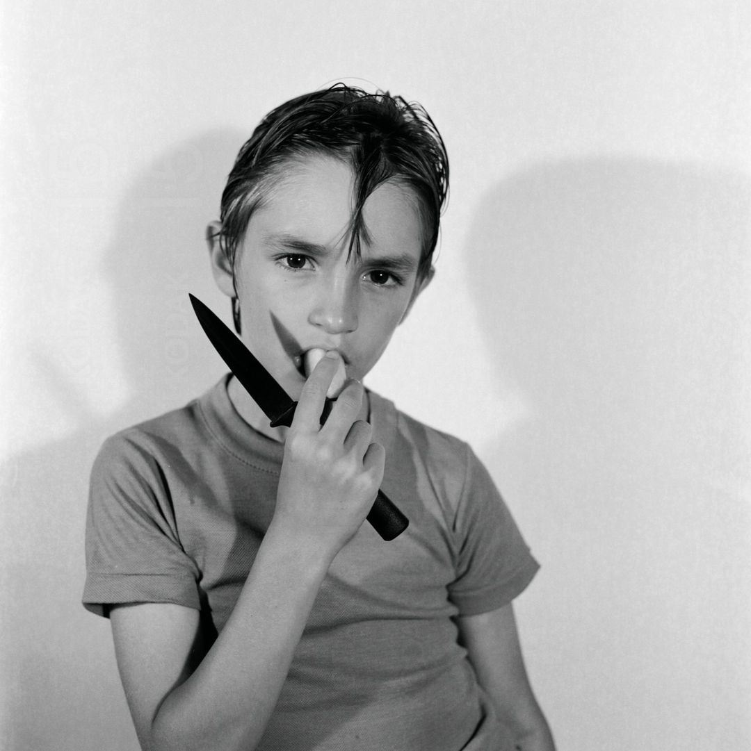 A young boy with dark hair holds a knife to his mouth in a black and white studio portrait with a plain white background.