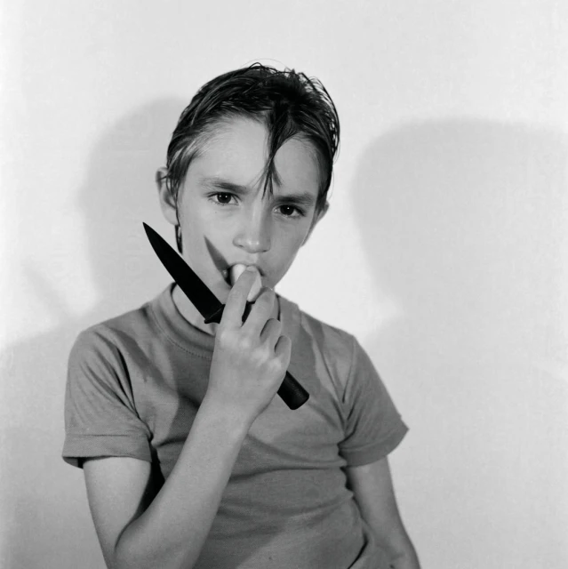 A young boy with dark hair holds a knife to his mouth in a black and white studio portrait with a plain white background.