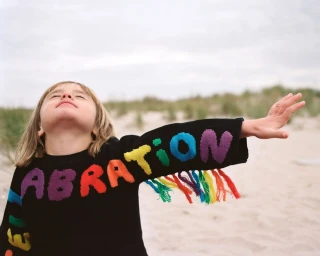 A young child wearing a black sweater with colorful knitted letters and fringe on the sleeve against a sandy beach background.