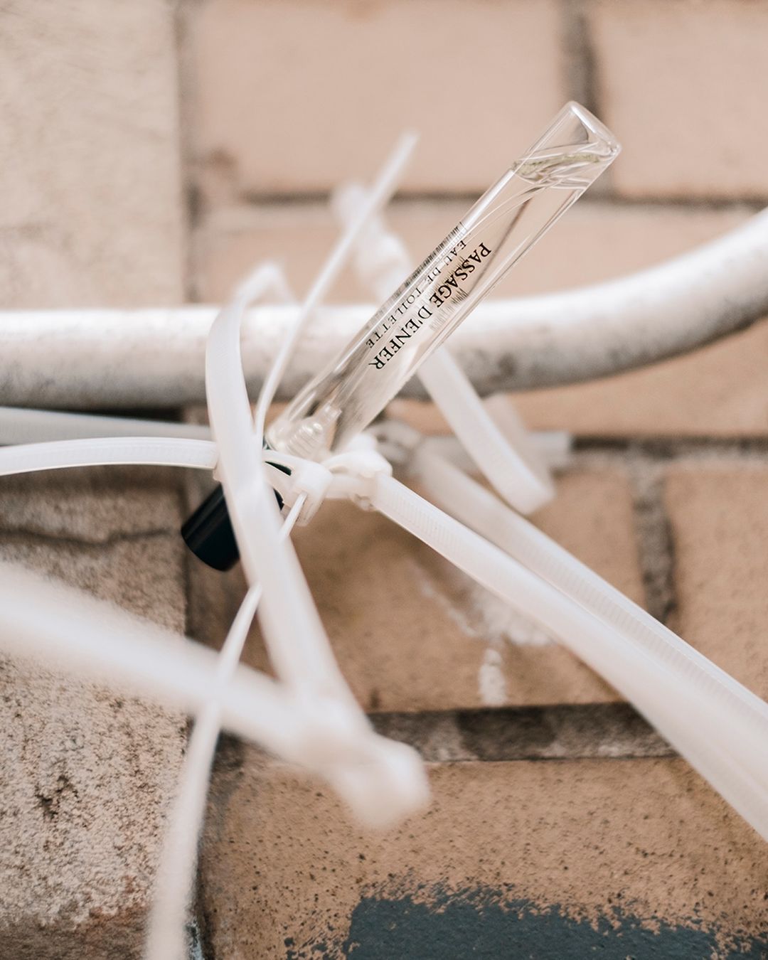 Perfume sample vial tied to a metal pipe against a brick wall, showcasing a minimalist presentation against an industrial backdrop.