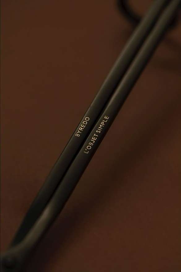Close-up shot of two black metal rods, branded with the names BYREDO and L'OBJET SIMPLE in gold lettering on a dark brown surface.