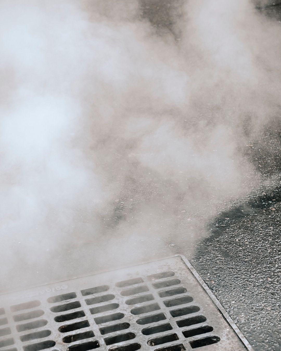 Steam rises from a metal grate in an urban street, creating a hazy effect in the outdoor environment, with a gray, muted color palette.