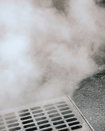 Steam rises from a metal grate in an urban street, creating a hazy effect in the outdoor environment, with a gray, muted color palette.