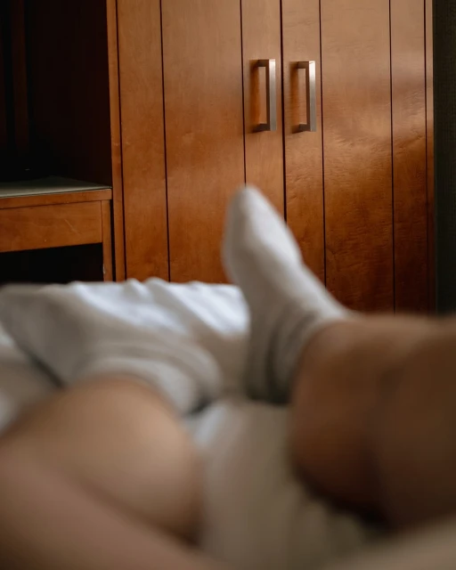 Close-up of an adult's legs and feet covered by a white bedsheet, with white socks, in a bedroom with a wooden closet in the background.