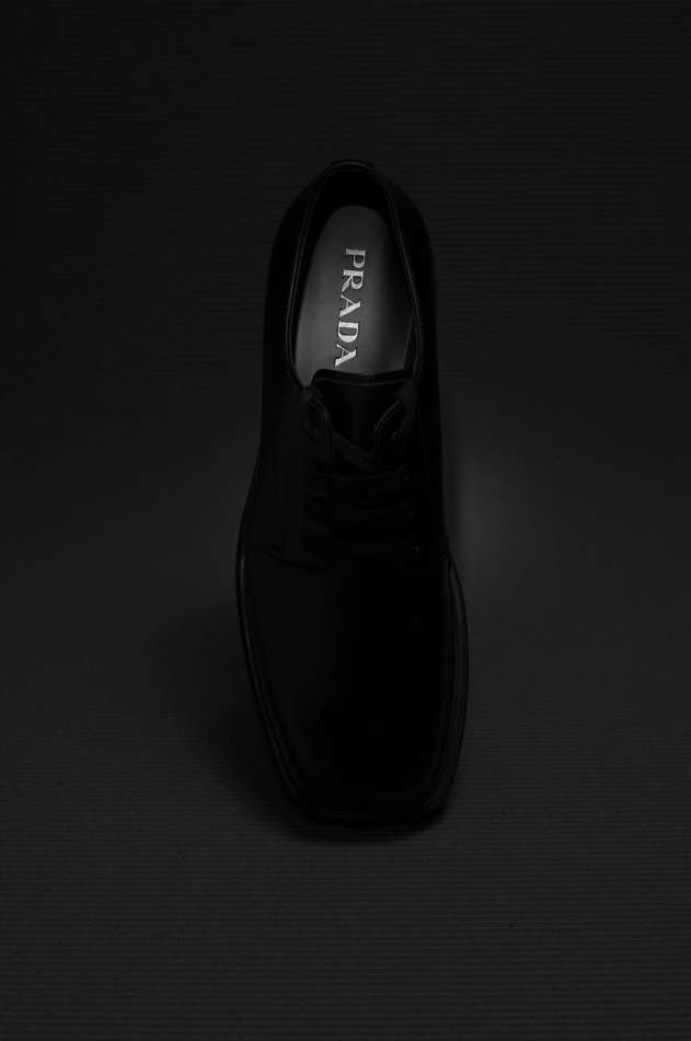 Black Prada leather shoe on a dark striped surface in a top-down studio shot, emphasizing texture and sleek design.