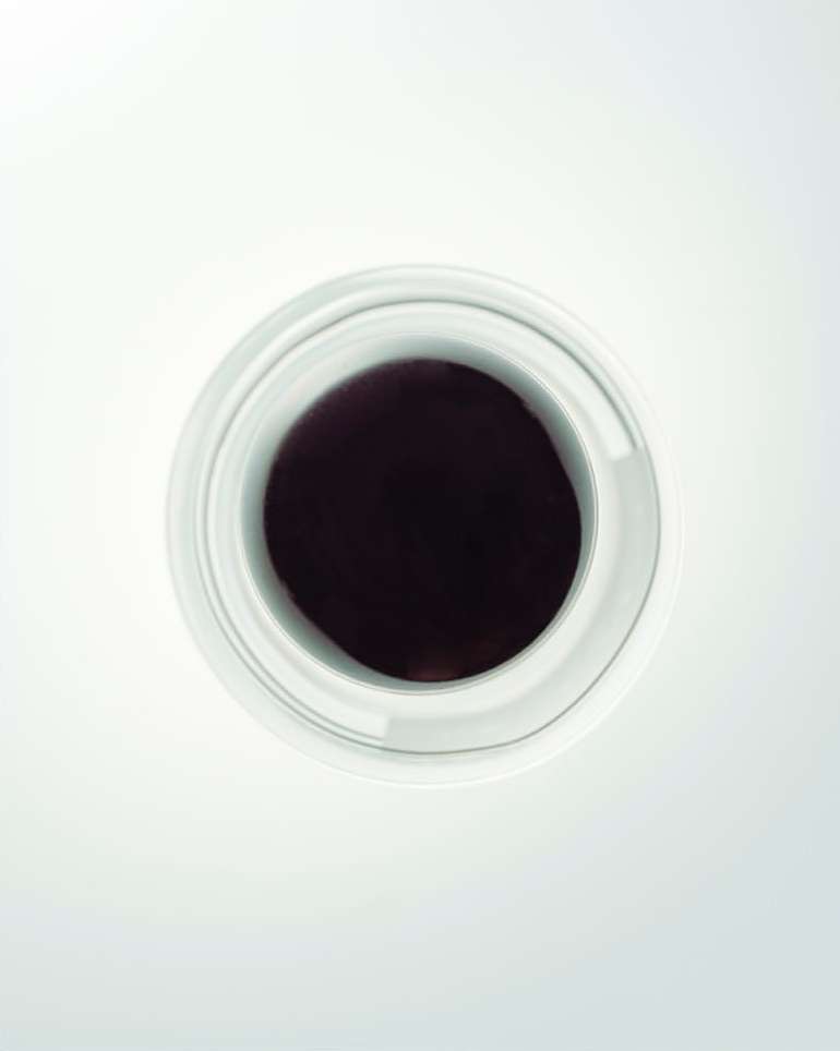 Overhead shot of a white ceramic container filled with dark liquid, possibly food or cosmetic product, on an illuminated white surface.