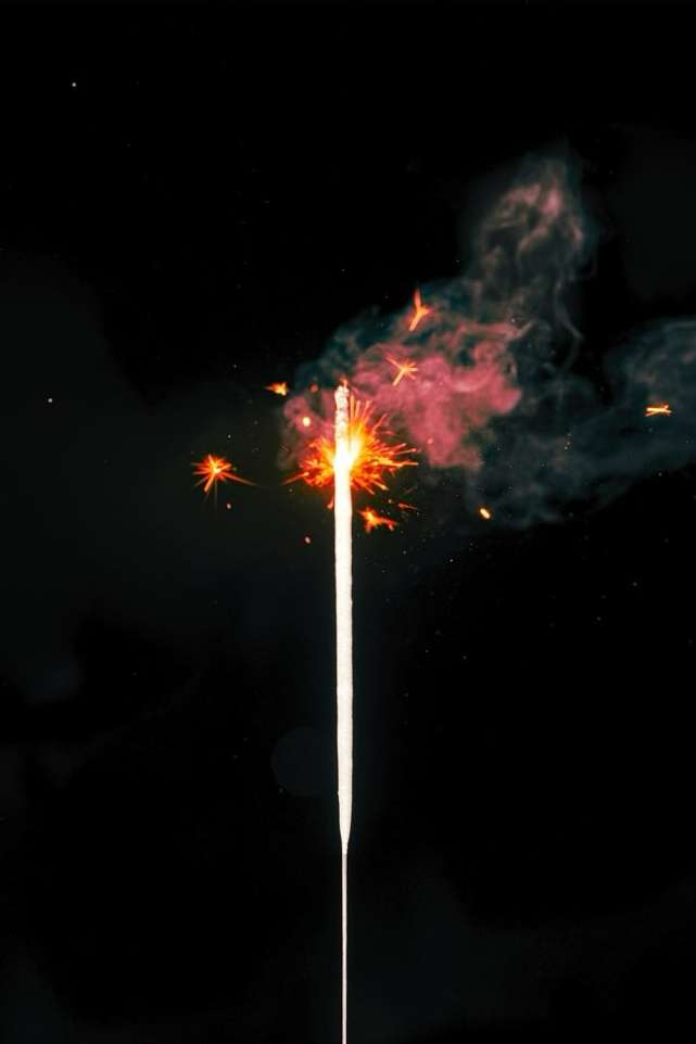 A lit sparkler emits bright sparks and smoke against a dark background, creating a vibrant contrast between light and shadow.